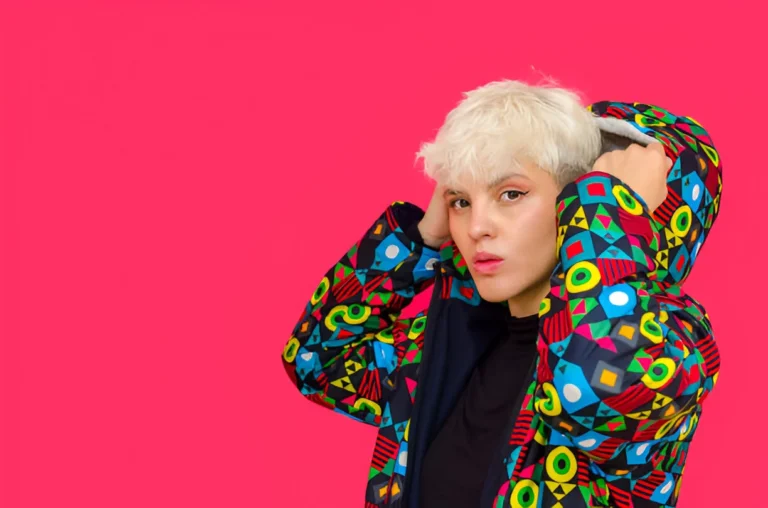 A person with short, platinum blonde bleached hair stands against a bright pink background, wearing a colorful geometric-patterned jacket with their hands adjusting the hood