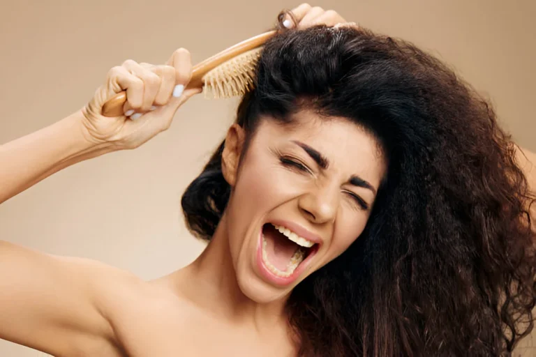 Woman with curly matted hair grimacing in pain while attempting to brush through tangled hair with a wooden brush against a neutral beige background