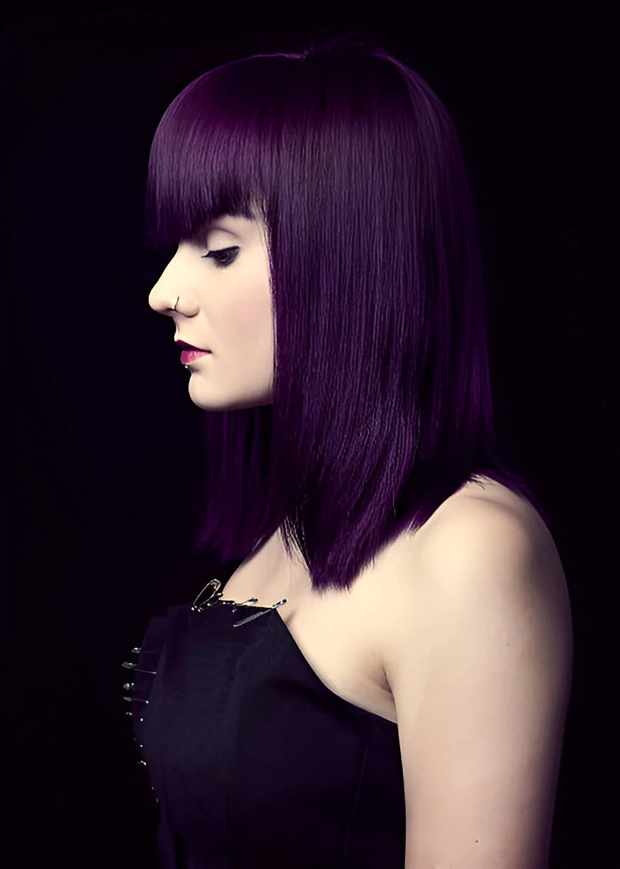A person with straight, dark purple hair with bangs, wearing a black strapless top with safety pin details, stands against a black background.