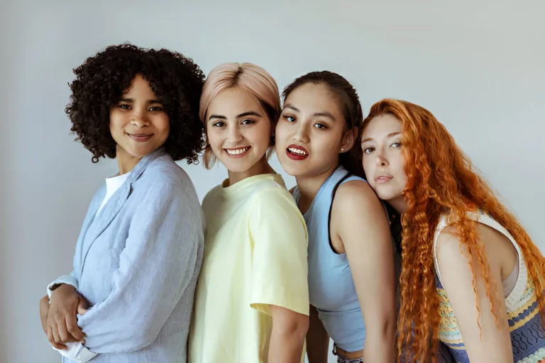 Four women with diverse hair textures and colors posing together against a light gray background, showcasing tight curls, straight blonde hair, sleek dark hair, and long wavy red hair.