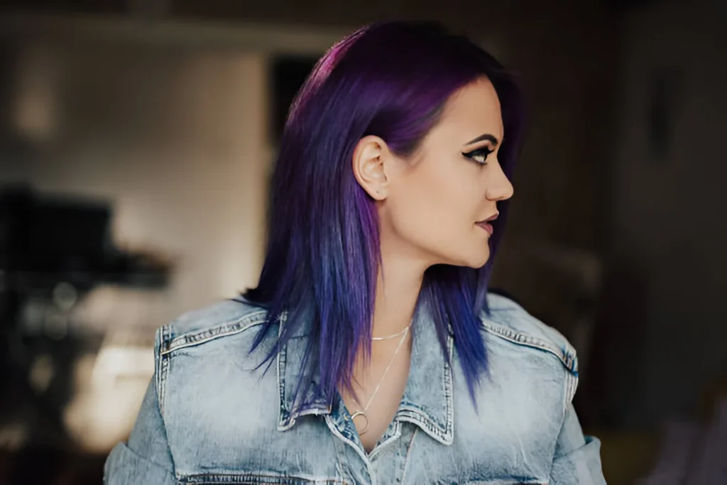A person with dark purple hair wearing a denim jacket and a delicate necklace stands indoors with a blurred background.