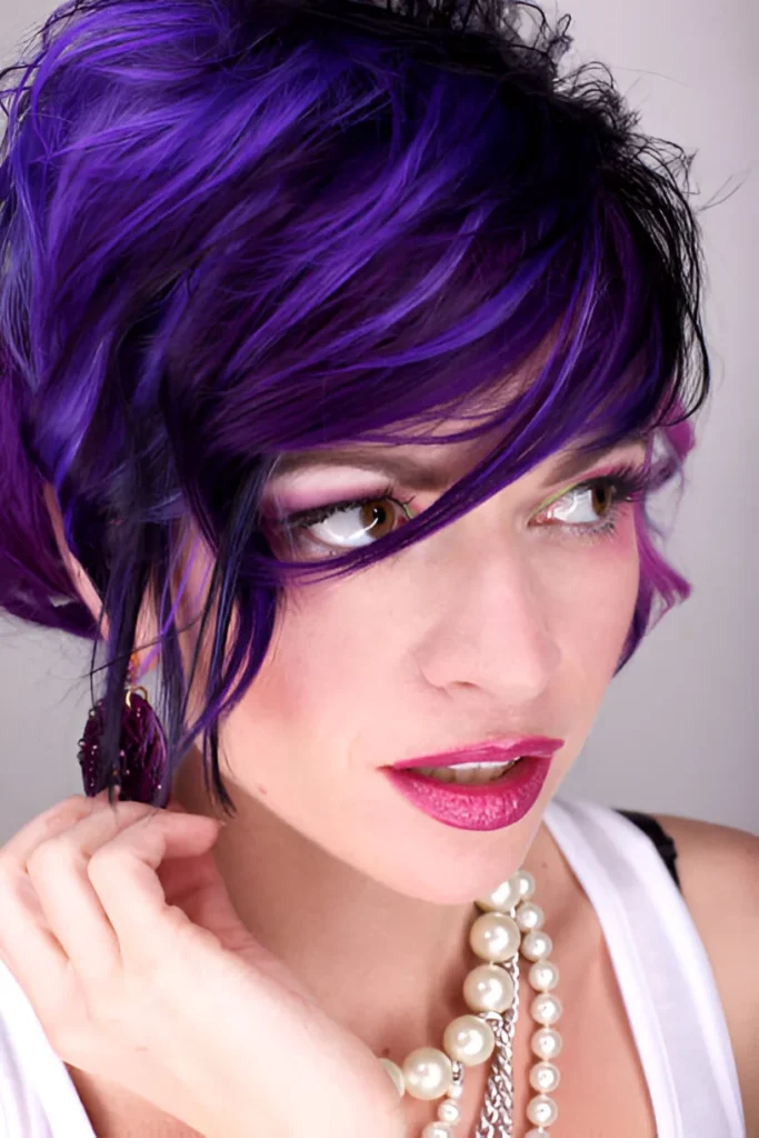 A person with vibrant dark purple hair styled in a tousled look, wearing a white top, a pearl necklace, and a matching purple earring against a plain background.