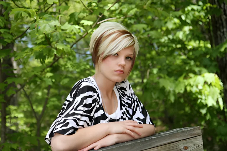Young woman with blonde and dark brown skunk hair featuring a short asymmetrical bob cut, wearing a zebra-print top, standing in front of lush green foliage.