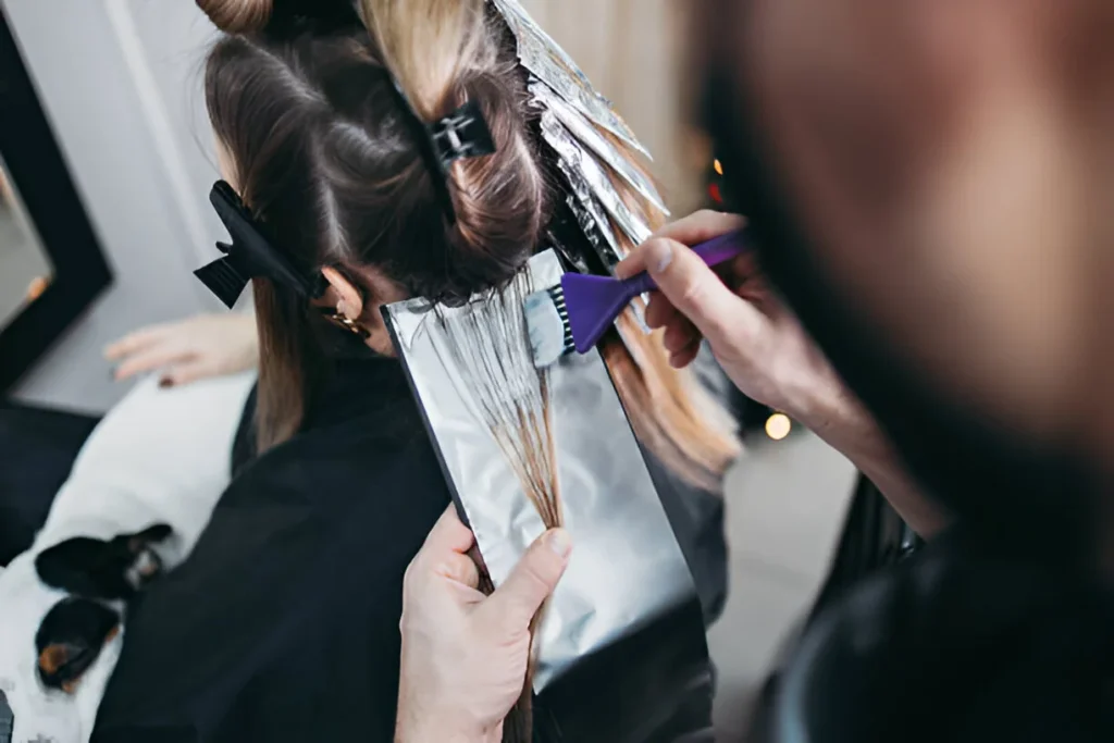 A stylist applies color to sections of hair wrapped in foil, using a purple brush, in a salon with a mirror and festive decorations in the background.
