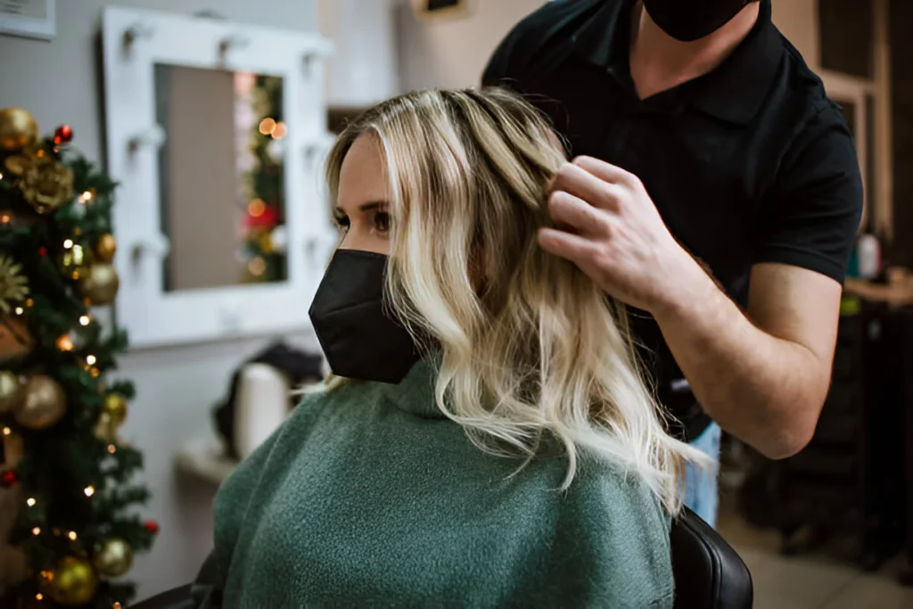 A stylist with blonde highlights on brown hair, wearing a green cape, is having their hair styled in a salon with a decorated Christmas tree and mirror in the background.