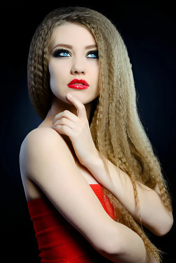 A person with long, crimped hair wearing a red strapless top poses against a dark background, with one hand resting on their chin.