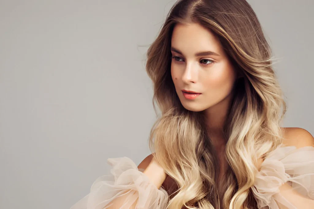 A person with long, wavy blonde hair wearing a sheer, ruffled dress against a light gray background.