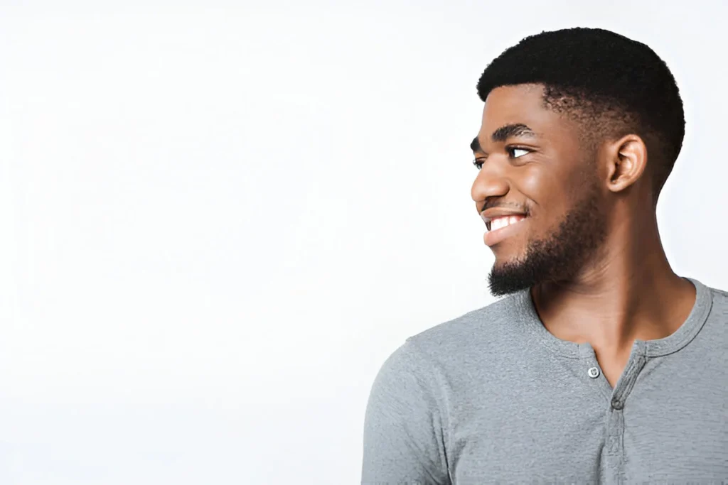 A person with a low taper fade curly hair style, wearing a gray shirt, smiling against a white background.