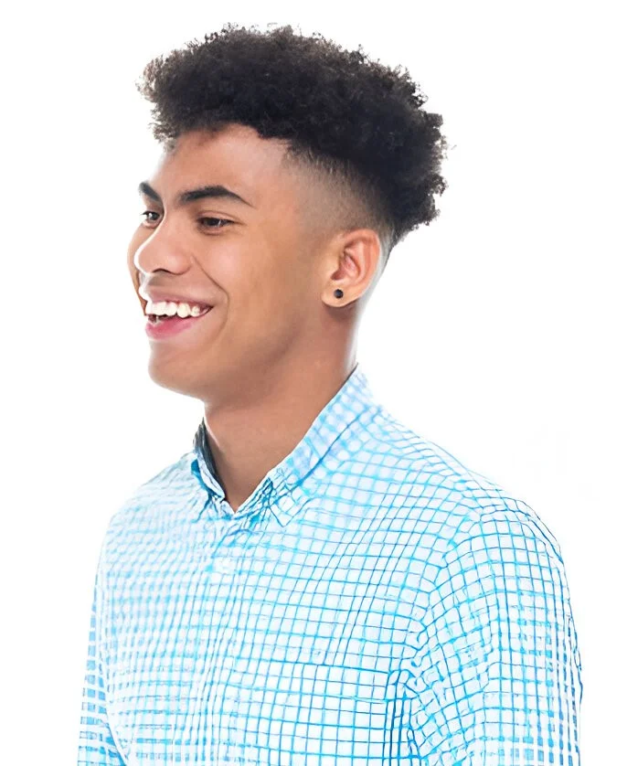 A person with a low taper fade curly hair style, smiling and wearing a blue checkered shirt against a white background.