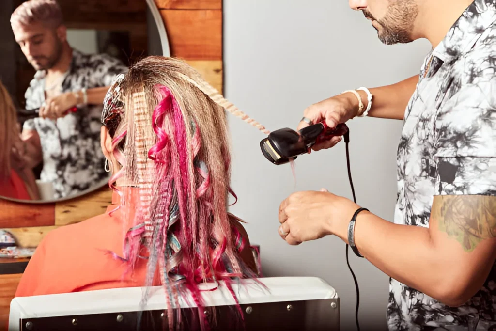 A stylist uses a crimping iron to style vibrant, multicolored hair with pink and blonde highlights in a salon setting, with another stylist working in the background.