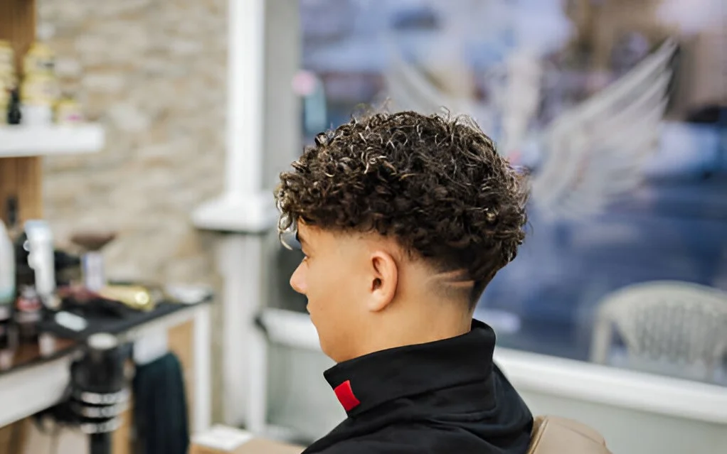 A person with a low taper fade curly hair style, sitting in a barbershop with a mirror and grooming products in the background.