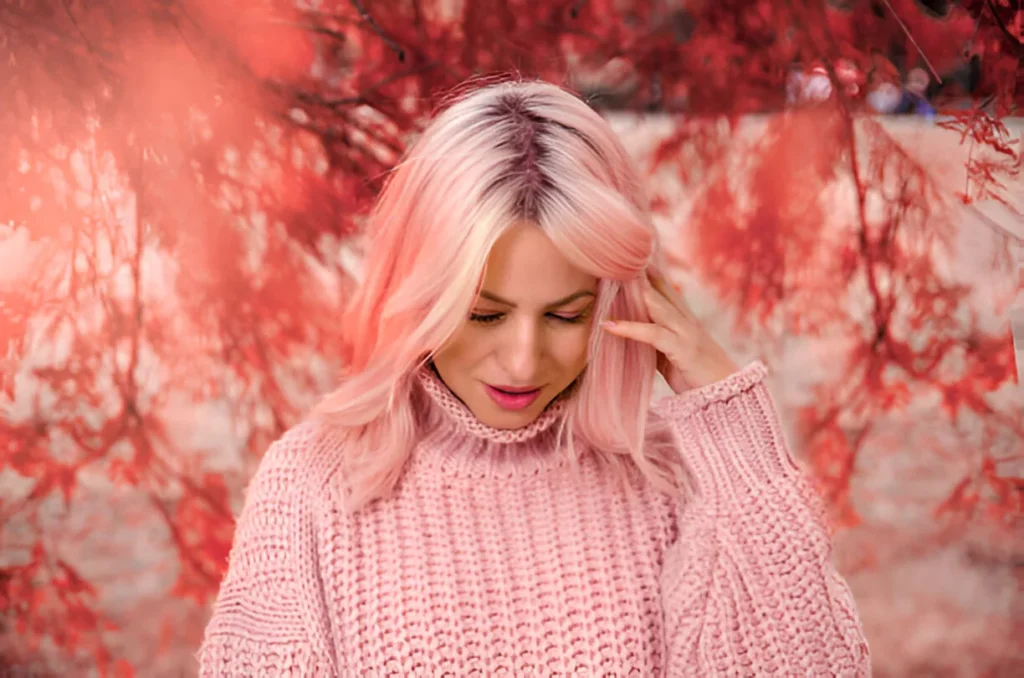 A person with rose gold hair wearing a matching pink sweater, standing with a hand to their head against a blurred red foliage background.