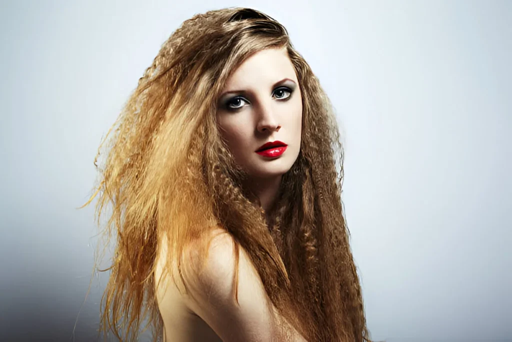 A person with long, voluminous crimped hair in shades of blonde and brown stands against a light gradient background, wearing a strapless top.