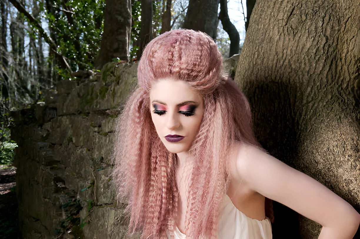 A person with long, pink crimped hair styled in an updo stands against a mossy stone wall and tree trunk in a forest setting, wearing a white strapless top.