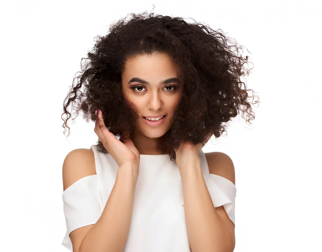 A person with voluminous curly hair, wearing a white off-shoulder top, smiling and holding their hair with both hands against a plain white background.