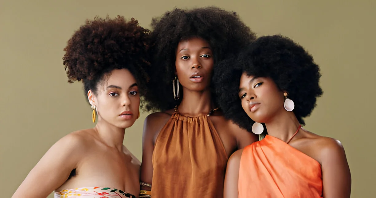 Three individuals with voluminous kinky hair posing together, wearing colorful sleeveless tops and large earrings against a neutral background.