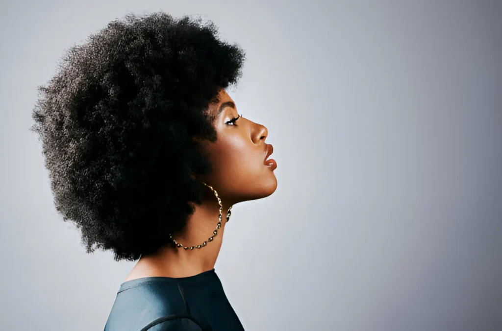 A person with voluminous kinky hair in profile, wearing large gold hoop earrings and a dark sleeveless top against a gray background.