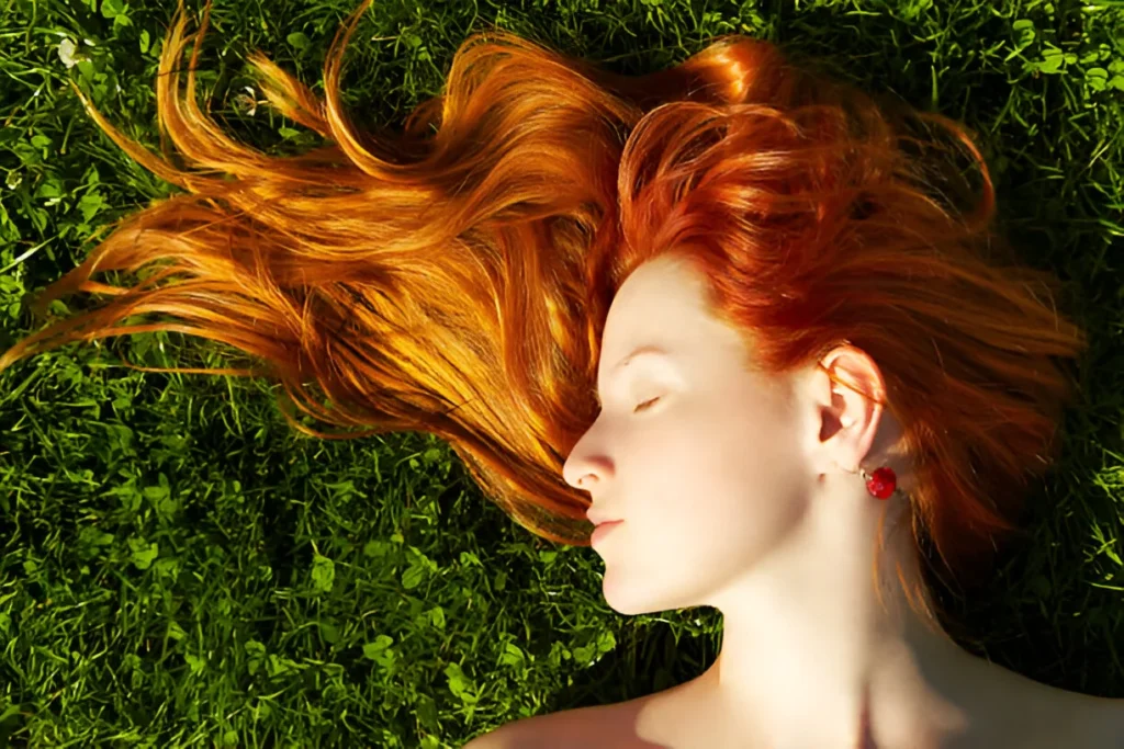 A person with long, flowing natural red hair lying on green grass, wearing red earrings, with eyes closed and a serene expression, captured in soft natural light.