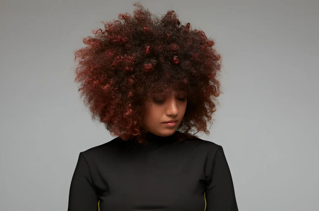 A person with voluminous curly hair dyed in shades of red and brown, wearing a black high-neck top with yellow accents, standing against a plain gray background.