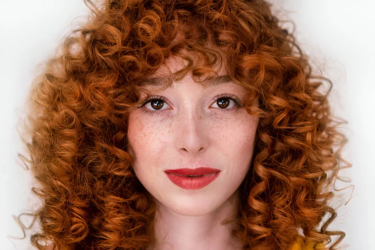 A person with voluminous natural red hair featuring tight curls, wearing a yellow top, with a confident expression, freckled skin, and bold red lipstick, set against a plain white background.