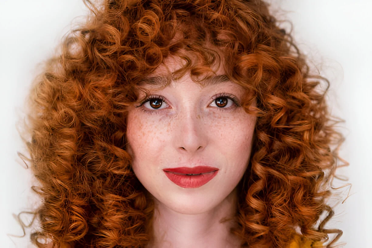 A person with voluminous natural red hair featuring tight curls, wearing a yellow top, with a confident expression, freckled skin, and bold red lipstick, set against a plain white background.