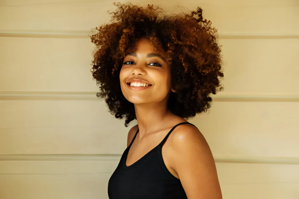 A person with voluminous kinky hair smiling against a light-colored background, wearing a black sleeveless top.
