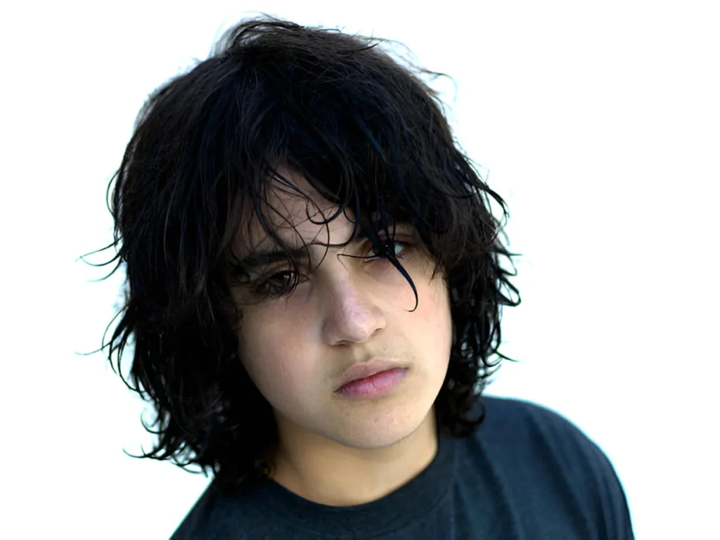 A person with tousled black wolf cut short hair against a white background, wearing a dark top.
